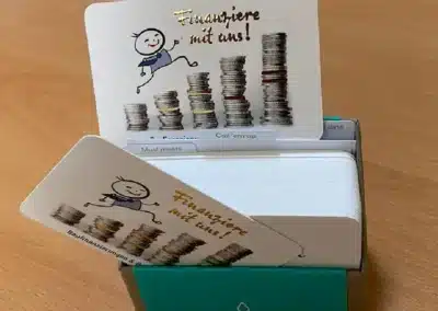 Eine Karteibox mit einem Stapel Visitenkarten, auf denen ein Strichmännchen abgebildet ist, das über Münzstapel springt, mit dem Text „Finanziere mit uns!“ auf einem Holztisch.