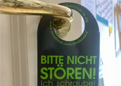 Auf dem Türhänger steht: „Bitte nicht stören! Ich schraube!“ was bedeutet: „Bitte nicht stören! Ich verarsche!“ auf Deutsch. Zusätzliche Übersetzungen und eine Website-URL sind sichtbar.