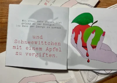 Aufgeschlagenes Heft mit einer deutschen Textseite und der Abbildung einer Hand, die einen roten Apfel mit grünem Blatt hält, aus dem eine rote Flüssigkeit tropft.