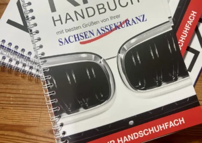 Spiralgebundene Autohandbücher, gestapelt auf einer Holzoberfläche. Auf dem Cover sind eine Nahaufnahme von Autoscheinwerfern und ein deutscher Text zu sehen, wobei das „KFZ Handbuch“ prominent hervorgehoben ist.