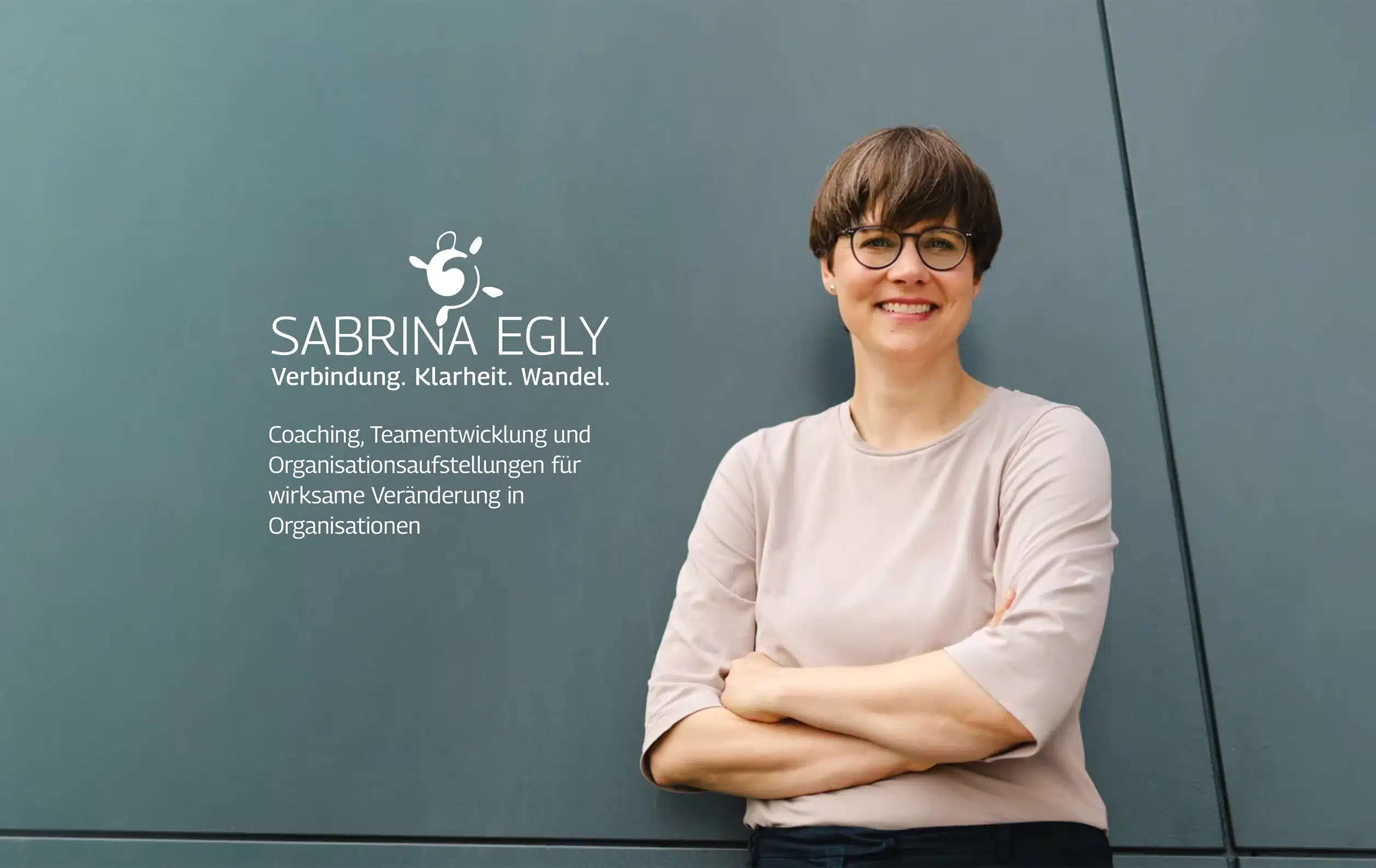 Sabrina Egly Eine Frau mit kurzen braunen Haaren, Brille und einem hellrosa Oberteil steht lächelnd mit verschränkten Armen vor einer grauen Wand. Neben ihr sind ein Logo und ein deutscher Text über Coaching, Teamentwicklung und organisatorische Veränderungen zu sehen.