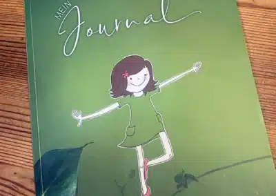 Ein grünes Tagebuch mit der Aufschrift "Mein Journal" auf dem Einband und der Illustration eines lächelnden Mädchens mit ausgestreckten Armen und rosa Schuhen. Die Ecken sind mit Blättern verziert und am unteren Rand steht "Zaza". Das Journal steht auf einer hölzernen Unterlage.
