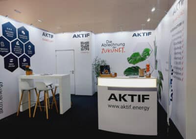 Ein moderner Messestand für AKTIF energy mit weißen Wänden, einer Theke, Barhockern und Infodisplays. Der Slogan "Die Abrechnung der Zukunft" und ein QR-Code sind sichtbar. Pflanzen und Broschüren schmücken die Theke.
