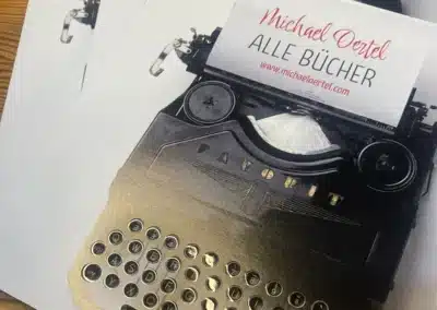 Eine Nahaufnahme von bedruckten Papieren mit einem alten Schreibmaschinenbild. Darüber befindet sich eine Visitenkarte von Michael Oertel mit der Aufschrift "ALLE BÜCHER" und einer Website-URL. Die Szene ist auf einer Holzfläche aufgebaut.